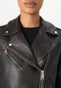 Giacca da motociclista in pelle nera con accessori in argento, chiusura con zip asimmetrica e colletto a scatto. Presenta una tasca con zip sul petto.