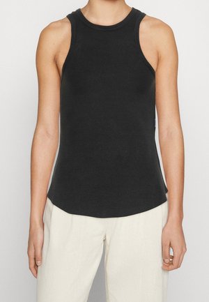 Czarny, prążkowany top bez rękawów noszony przez osobę z rozluźnionymi ramionami, zestawiony z luźnymi białymi spodniami na neutralnym tle.