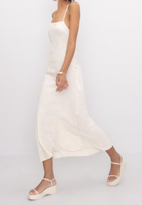 Robe midi crème avec des fines bretelles, présentant un motif animalier subtil. Associée à des sandales à plateforme beiges. Texture lisse.