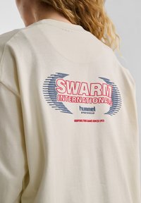 Beige Sweatshirt mit langen Ärmeln, das auf dem Rücken einen großen Grafikdruck zeigt, mit "SWARM INTERNATIONALS" in rotem und blauem Text.
