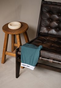 Tyrkysové šortky s bílým lemem, na kterém je nápis "ŠŤASTNÉ ŠORTKY", přehnuté přes hnědou koženou židli vedle dřevěného stolu s kulatou kamennou miskou.