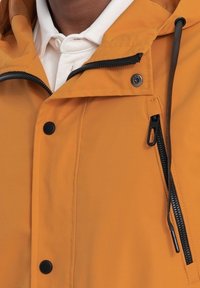 Veste orange résistante à l'eau avec une fermeture éclair et des boutons-pression, dotée d'accents noirs et de cordons ajustables à la capuche.