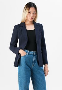 Mladá žena s dlouhými vlasy má na sobě navy sako přes černý top a volné modré džíny, stojí s jednou rukou v kapse na bílém pozadí.