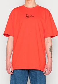 Oranžové oversized tričko z bavlny, s černým signaturním logem na hrudi a krátkými, širokými rukávy.