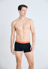 Sorte boxer shorts med en orange "TOMMY HILFIGER" talje, der har et tætsiddende design og glat stof, modeleret på en overlæsset mandlig figur.