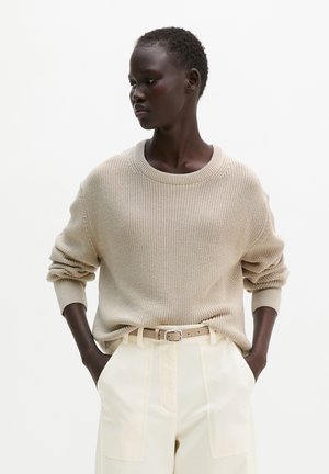 Femme aux cheveux courts portant un pull en maille beige rentré dans un pantalon crème taille haute avec une ceinture beige, mains dans les poches, regardant sur le côté.