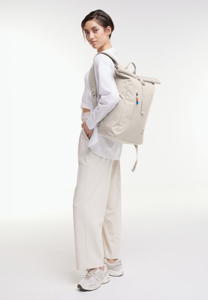 Nuori nainen vaaleassa rentossa asussa, jolla on beige rullattava reppu, seisoo sivuttain ja käsi taskussa valkoista taustaa vasten.