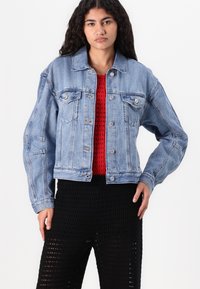 Hele sinine denim-jakk klassikalise lõikega, kahe rinnatasku, hõbedaste nuppudega ja nähtavate õmblusdetailidega, kantud punase kudumipealse peal.