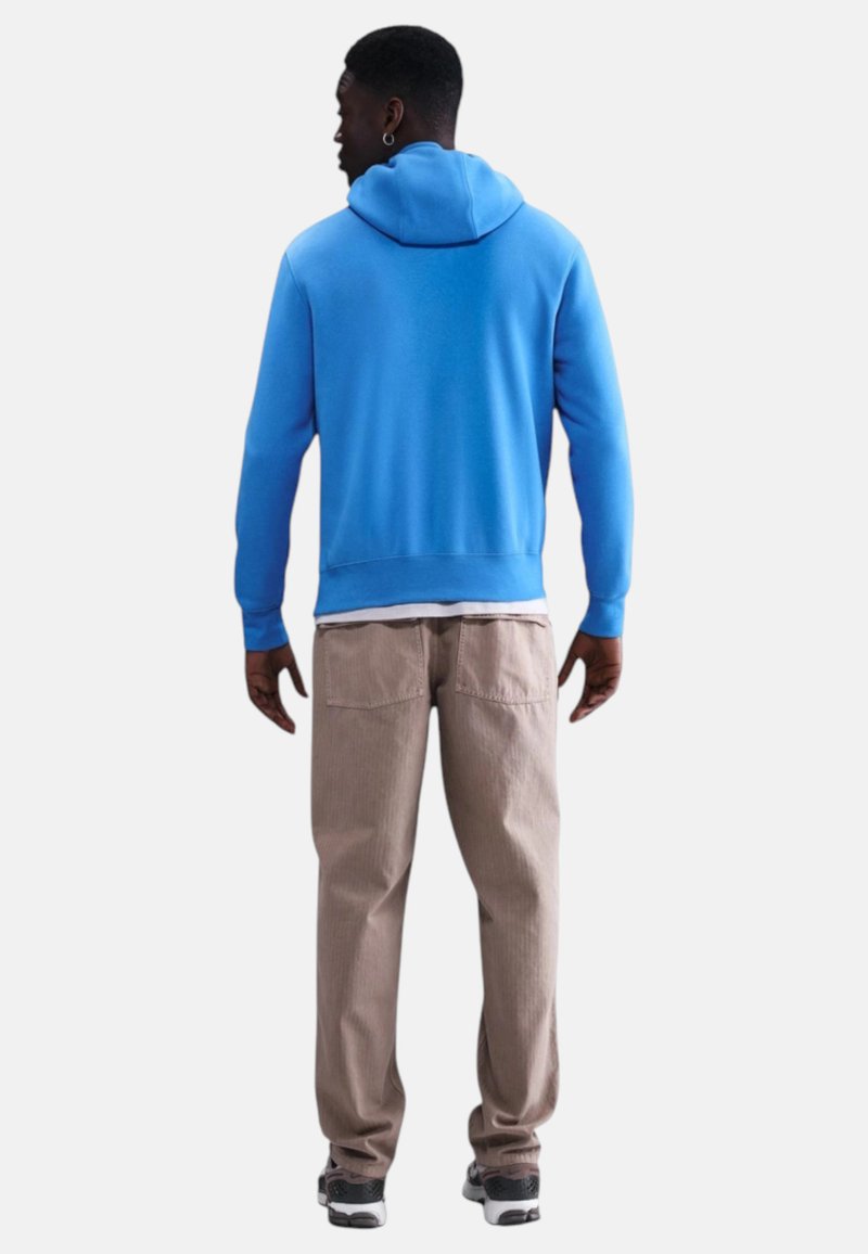 Uomo che indossa una felpa con cappuccio azzurra brillante, pantaloni beige, sneakers nere e bianche, in piedi e di spalle su uno sfondo semplice.