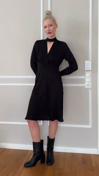 Γυναίκα με ξανθά μαλλιά μαζεμένα σε κότσο, που φοράει μαύρο φόρεμα με μακριά μανίκια και άνοιγμα στο ντεκολτέ, καθώς και μαύρα μποτάκια μέχρι τον αστράγαλο, σε εσωτερικό χώρο.