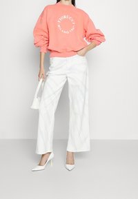 Koraalroze sweatshirt met logo, witte wijde broek met subtiele marmering, en witte puntige hakken met neonaccenten.