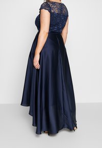 Navy sininen iltapuku pitsihihnoilla, istuva yläosa ja valuva satiinihame. Sisältää hieman kohotetun takahameen ja sileän pinnan.