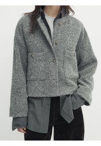 Veste cropped en laine grise texturée avec deux poches avant et fermeture à boutons ; superposée sur une chemise sombre à boutons et un pantalon sombre.
