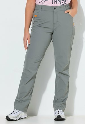 Persoon die grijze outdoorbroek met zakken draagt, witte en zwarte sneakers, staande tegen een witte muur, met één hand in de zak.