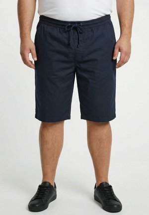 Mand iført marineblå snørebukser, hvid t-shirt og sorte sneakers, stående foran en ensfarvet lys baggrund.