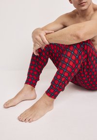 Rode geruite loungebroek met blauwe accenten, elastische boorden en een zachte, gestructureerde stof. Geplaatst op een kale vloer met blote voeten zichtbaar.