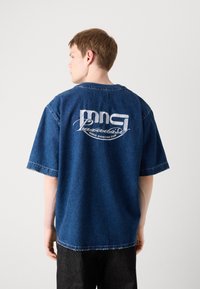 Blå denim oversized t-skjorte med brodert hvit tekst på baksiden, med rund hals og korte ermer. Uferdig kantdetalj.