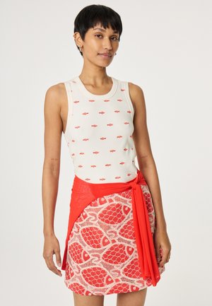 Junge Frau trägt weißes ärmelloses Oberteil mit kleinen roten Fischmustern und Rock mit roten Fischmustern und gebundenem rotem Gürtel.