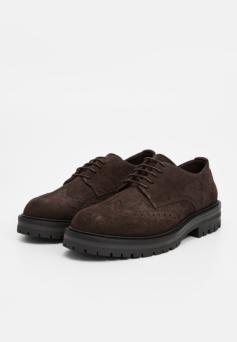 Para ciemno-brązowych zamszowych butów brogues z czarnymi grubymi gumowymi podeszwami i sznurowadłami na białym tle.
