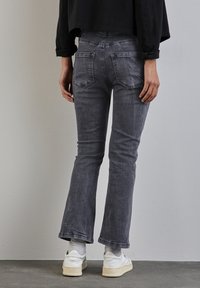 Persona vestida con jeans negros desgastados, zapatillas blancas y calcetines grises, de pie frente a una pared y un suelo gris claro liso.