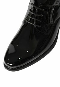 Scarpa elegante in pelle nera lucidata con lacci e cuciture dettagliate su sfondo bianco.