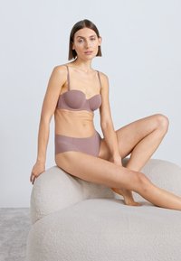 Svetlomauve podprsenka s tenkými ramienkami a tvarovanými košíčkami, v kombinácii s rovnako sfarbenými vysokým pásom nohavičiek, sediaca na textúrovanom, zaoblenom kresle.
