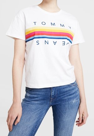 Camiseta blanca de manga corta con el logotipo "TOMMY", que presenta rayas horizontales en amarillo, rosa y azul a lo largo del pecho. Corte casual.