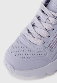 Primo piano di una scarpa da ginnastica lavanda con dettagli perforati, lacci spessi intrecciati e una suola strutturata su sfondo bianco.