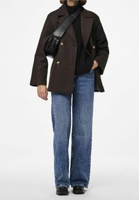 Brun dubbelknäppt blazer, svart polotröja, blå vida jeans och svarta skor. Blazern har guldknappar och en avslappnad passform.