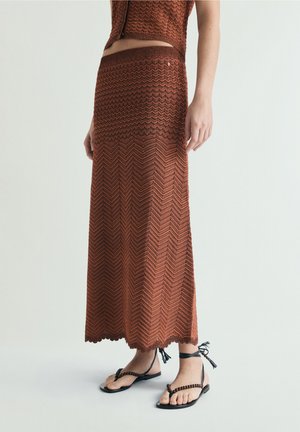 Mujer con falda maxi de punto marrón con estampado y top corto a juego, combinados con sandalias negras atadas al tobillo y con cuentas.