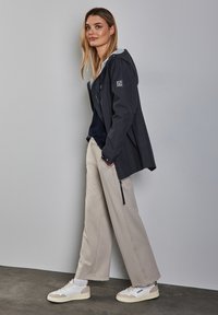 Veste navy avec capuche, doublure grise, coupe spacieuse. Pantalons beige clair avec fermetures éclair sur les côtés. Baskets blanches avec des accents beiges.