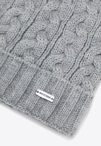 Šedá pletená čepice s copánkovým vzorem a žebrovaným lemem. Na boku je stříbrná značka s nápisem "WITTCHEN". Měkká struktura.