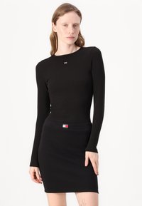 Svart ribbestrikket langermet topp med rund hals, kombinert med et tettsittende svart skjørt med logobånd i midjen. Glatt tekstur, minimalistisk design.