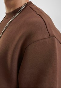 Sudadera marrón con un acabado texturizado, que presenta un escote de cuello redondo y una costura de hombro prominente. Se puede ver un collar de cadena plateada.
