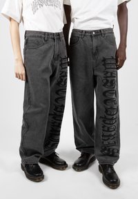 Baggy mörkgrå jeans har ett framträdande svart grafiskt tryck längs vänster ben. Kombineras med svarta snörskor och enfärgade vita t-shirts.