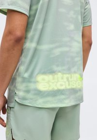 Άτομο που φοράει αθλητικά ρούχα ανοιχτού πράσινου χρώματος με κοντά μανίκια και σορτς, κείμενο «outrun excuses» σε φωτεινό κίτρινο κοντά στο κάτω μέρος του πουκαμίσου.