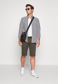 Camisa listrada em preto e branco, t-shirt branca, calções verde-oliva e ténis brancos de cano alto. Mala de ombro preta com detalhes coloridos.