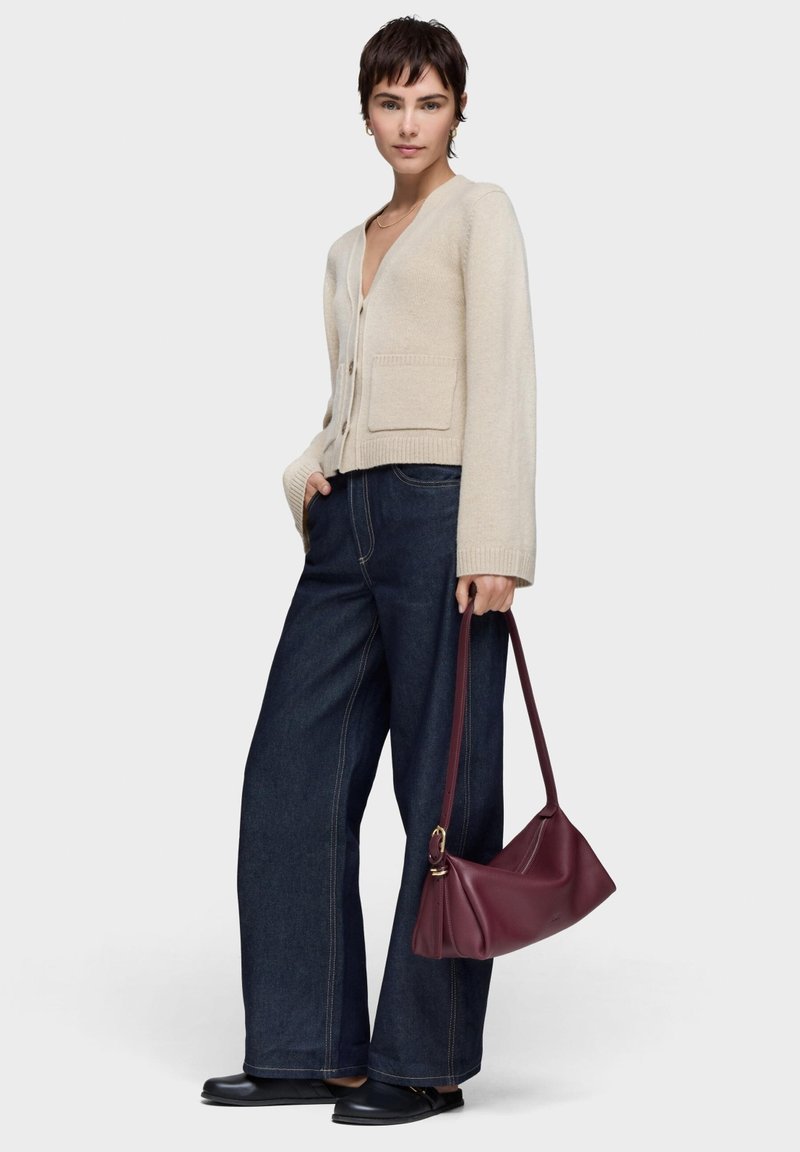 Cárdigan beige con escote en V y bolsillos frontales, combinado con pantalones de mezclilla oscuros de pierna ancha. Bolso de hombro burdeos con forma curva y detalles en dorado.