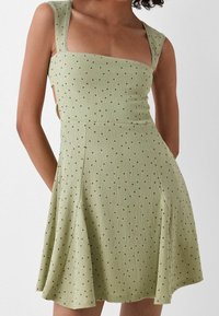 Abito a coste verde chiaro con scollatura quadrata, spalline larghe, tagli laterali e piccoli motivi a pois neri. L'orlo a pieghe della gonna aggiunge movimento.