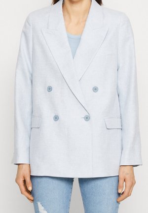 Kvinde iført en lyseblå dobbeltradet blazer over en blå top og revnede lyseblå jeans, stående mod en hvid baggrund.