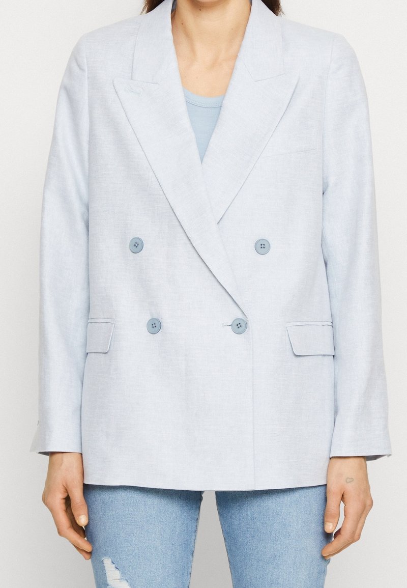 Femme portant un blazer croisé bleu clair sur un haut bleu et un jean déchiré bleu clair, debout sur un fond blanc.