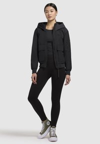 Veste à capuche noire avec deux poches avant, fermeture éclair et poignets élastiqués. Portée sur un haut noir et des leggings noirs, avec des baskets montantes vertes.