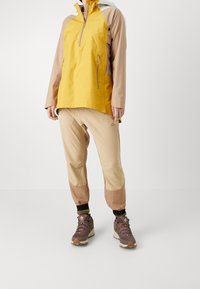 Gul och beige utomhusjacka med dragkedja, parat med beige byxor med bruna detaljer och bruna vandringskängor.