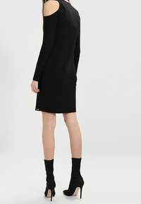 Svart långärmad klänning med öppningar på axlarna, figurnära snitt, knälång, slät yta och metallknappdetalj på sidan.
