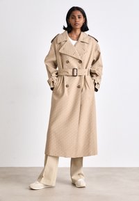Manteau trench à motif beige avec fermeture croisée, taille ceinturée et épaulettes; associé à des pantalons crème et des chaussures.