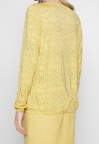 Langärmlige gelbe Bluse mit kariertem Muster, mit elastischen Bündchen und lockerer Passform. Der Stoff hat eine glatte Textur und fällt sanft.