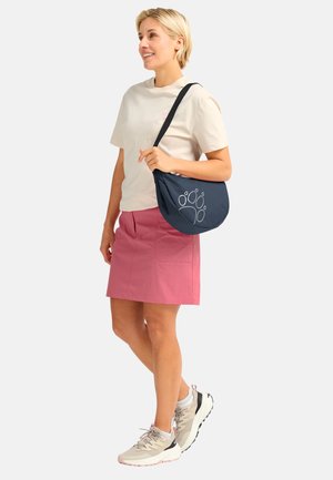 Vrouw met beige T-shirt, roze rok, beige sneakers, draagt een marineblauwe schoudertas met een pootafdrukontwerp, staat en lacht opzij.