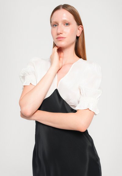 Zwarte satijnen jurk met een witte top met pofmouwen, voorzien van een diepe V-hals en smockmanchetten. Gladde, contrasterende stoffen met strakke lijnen.