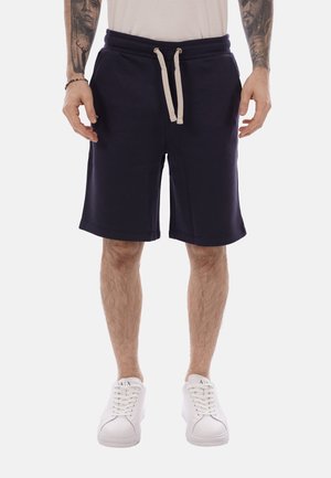 Uomo che indossa pantaloncini con coulisse blu navy, scarpe da ginnastica bianche e una camicia bianca, in piedi con le braccia rilassate lungo i fianchi.