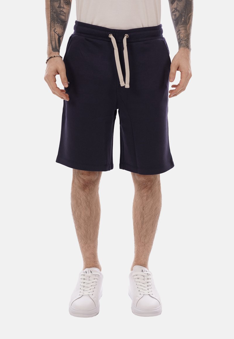 Homme portant un short bleu marine à cordon, des baskets blanches et une chemise blanche, debout les bras détendus le long du corps.